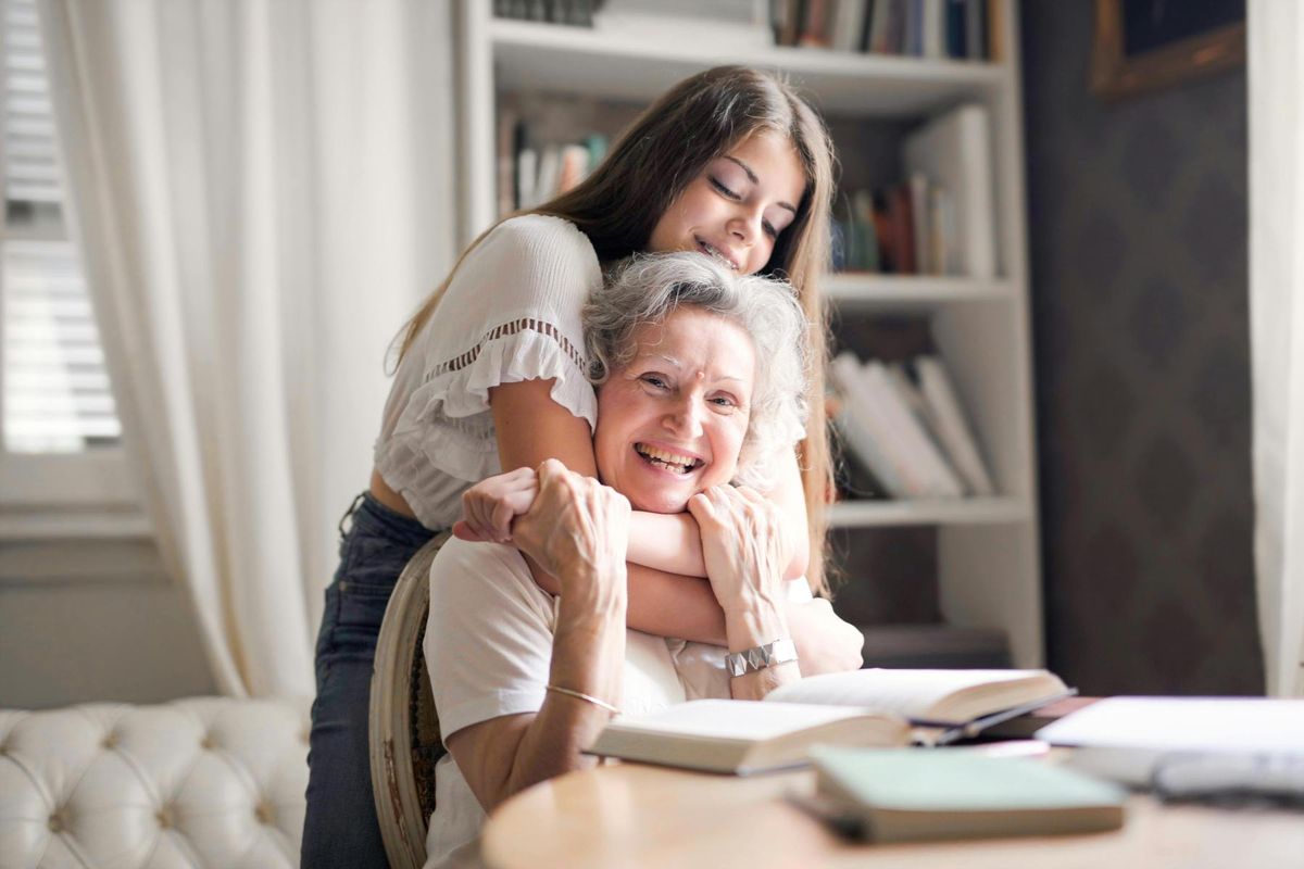 A young companion hugs a delighted older woman at her reading desk